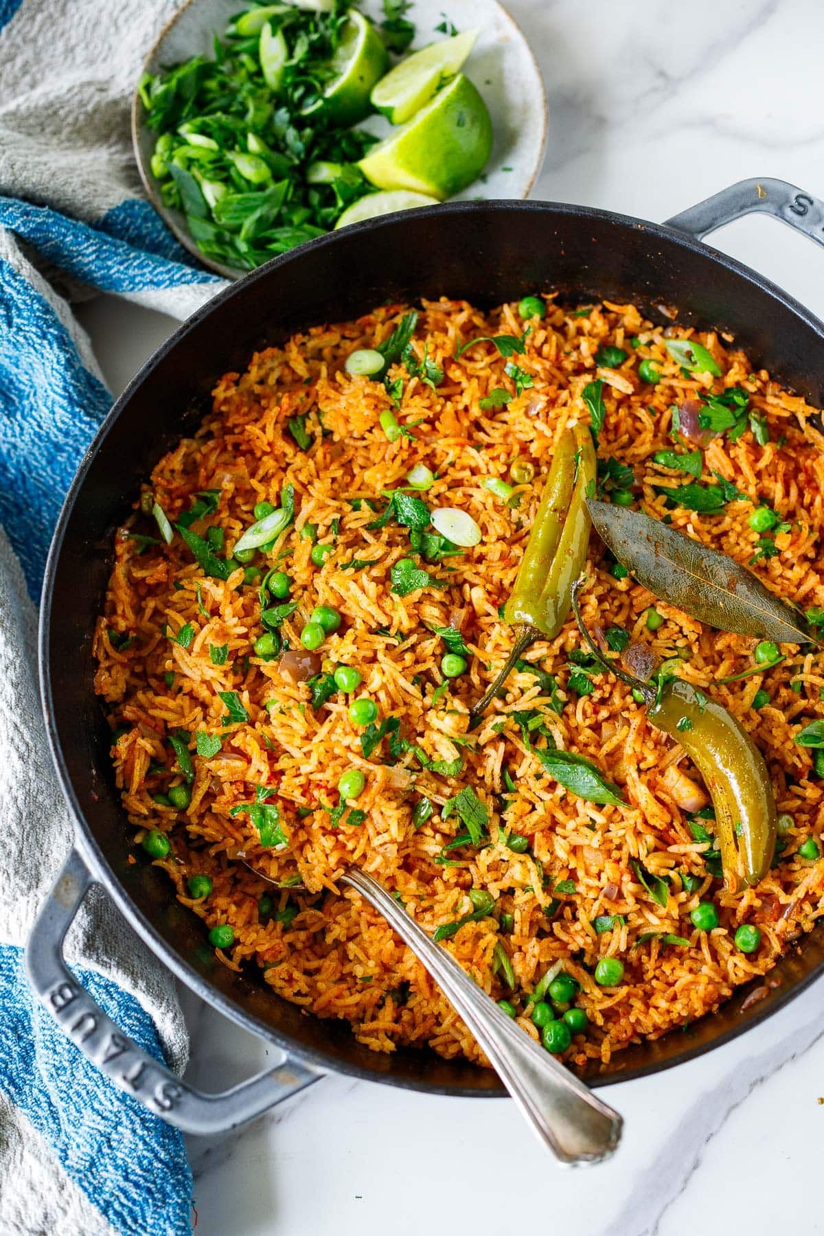 Spanish rice in a pan with peas, green onions, cilantro, bay leaf, serrano chilies, and a serving spoon. 