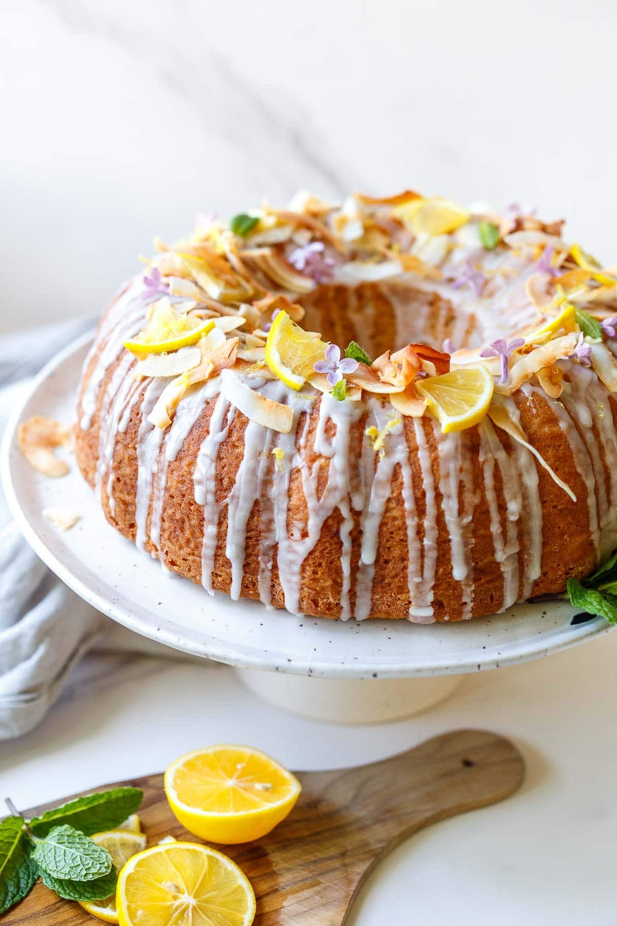 vegan lemon bundt cake with lemon glaze, lemon quarters, purple flowers, toasted coconut, lemon zest, and mint, on cake plate.