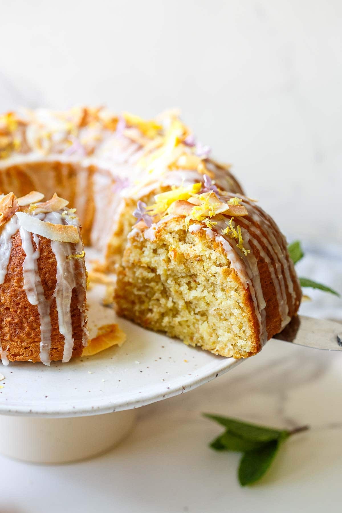 spatula removing a slice of vegan lemon bundt cake with lemon glaze, lemon zest, toasted coconut, and purple flowers from cake plate.