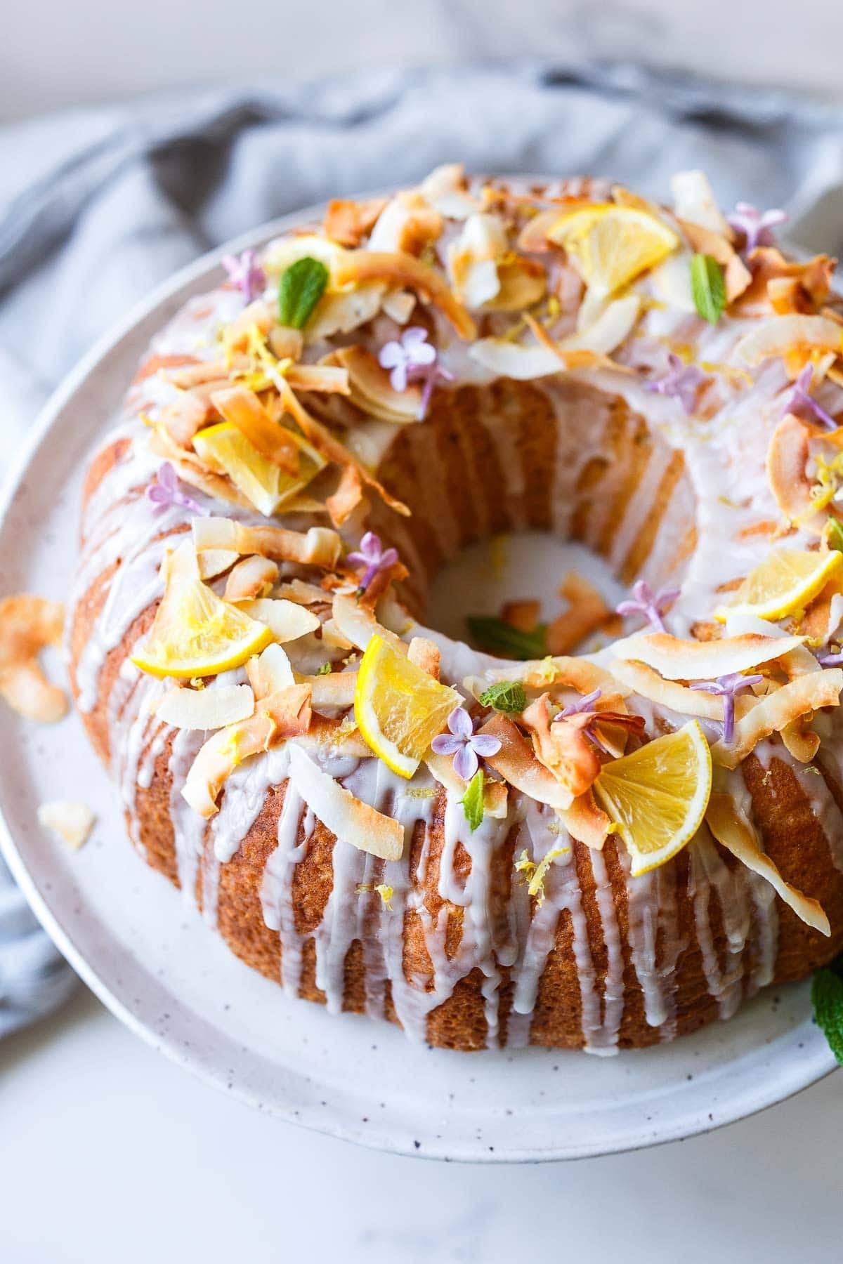 vegan lemon bundt cake with lemon glaze drizzled over top and garnished with lemon quarters, toasted coconut, edible flowers, and fresh mint. 