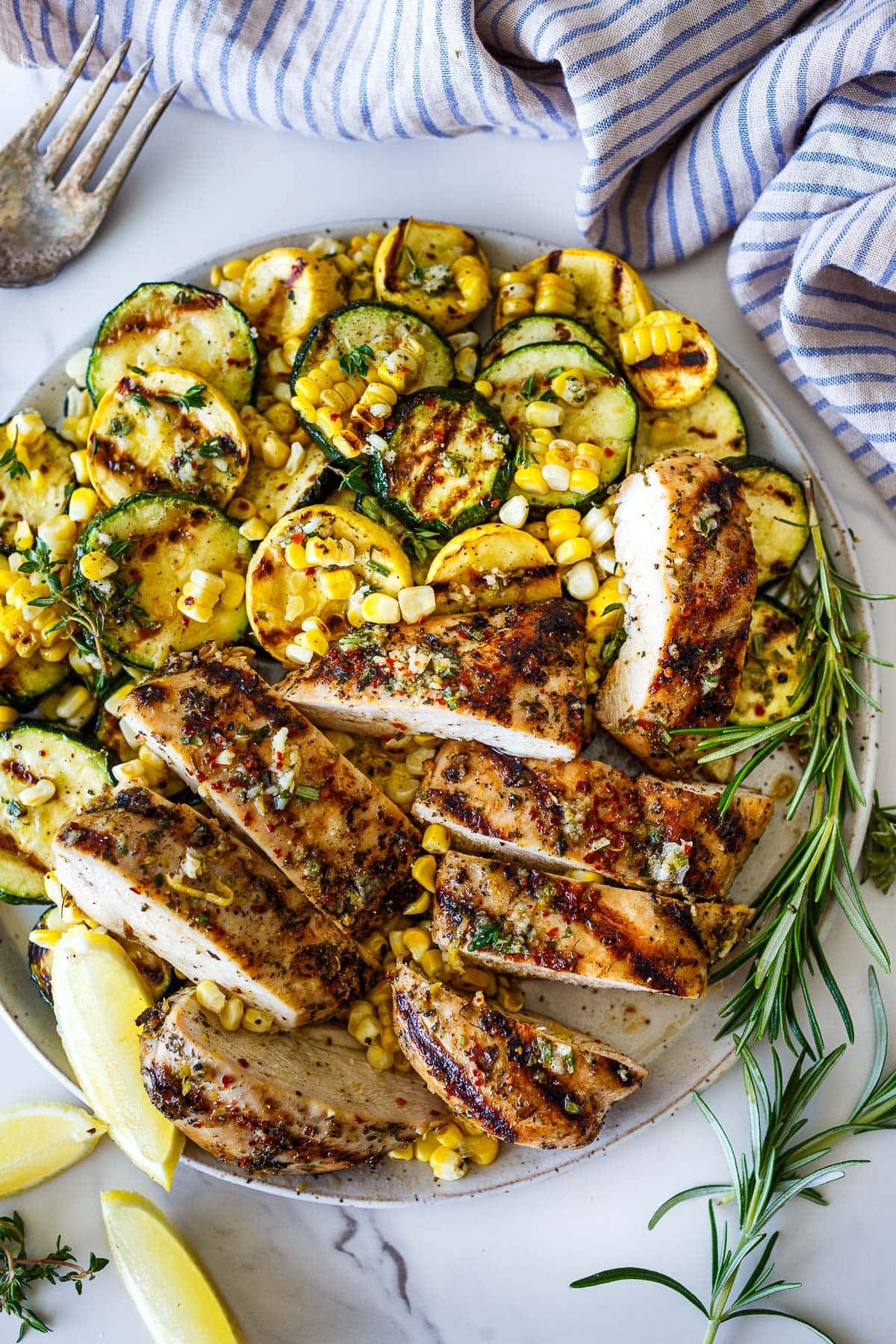 platter of Mediterranean grilled chicken, sliced, with grilled corn and zucchini, topped with fresh herbs.