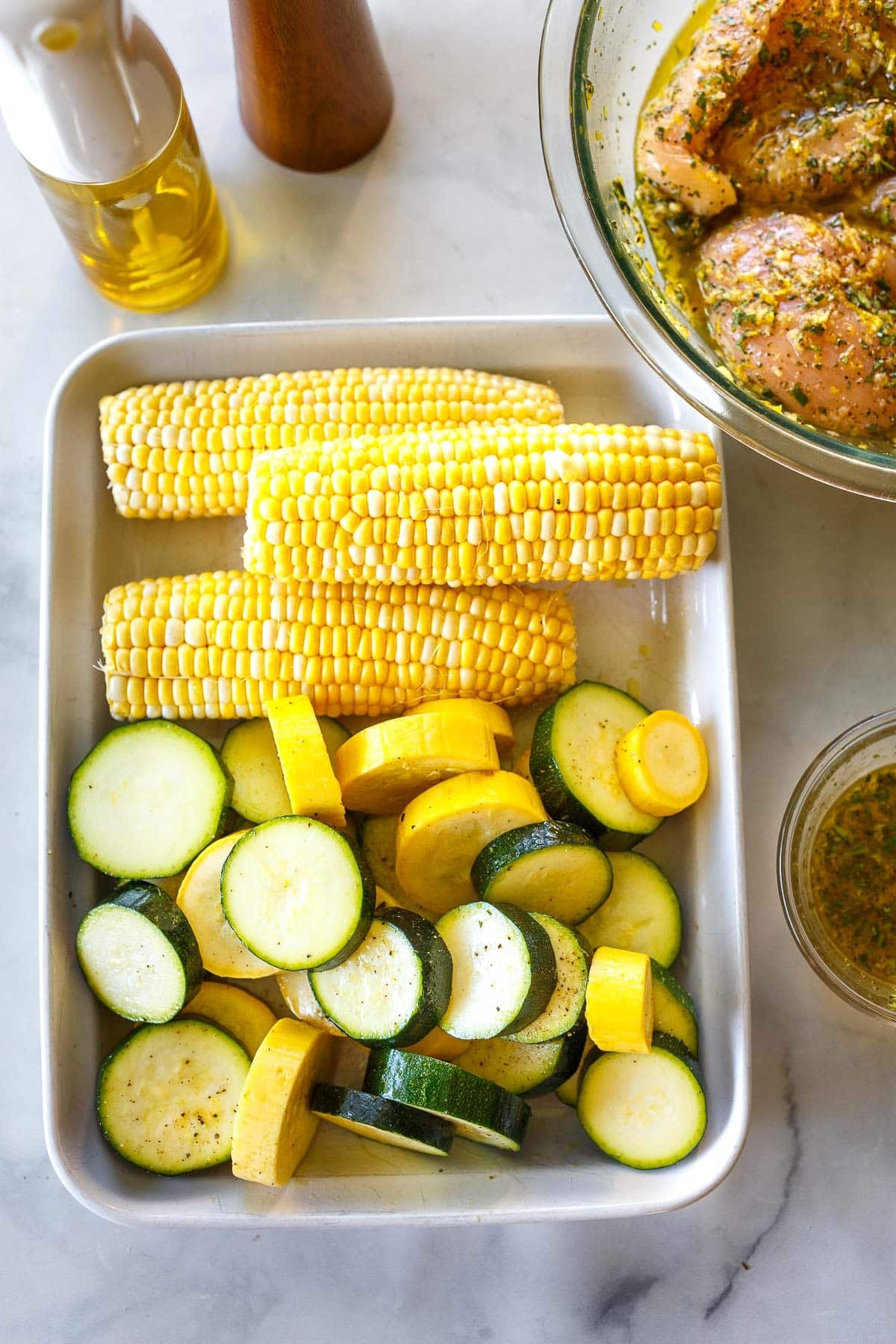 rectangle baking dish with three ears of corn next to sliced zucchini and yellow squash tossed with olive oil, salt, and black pepper.