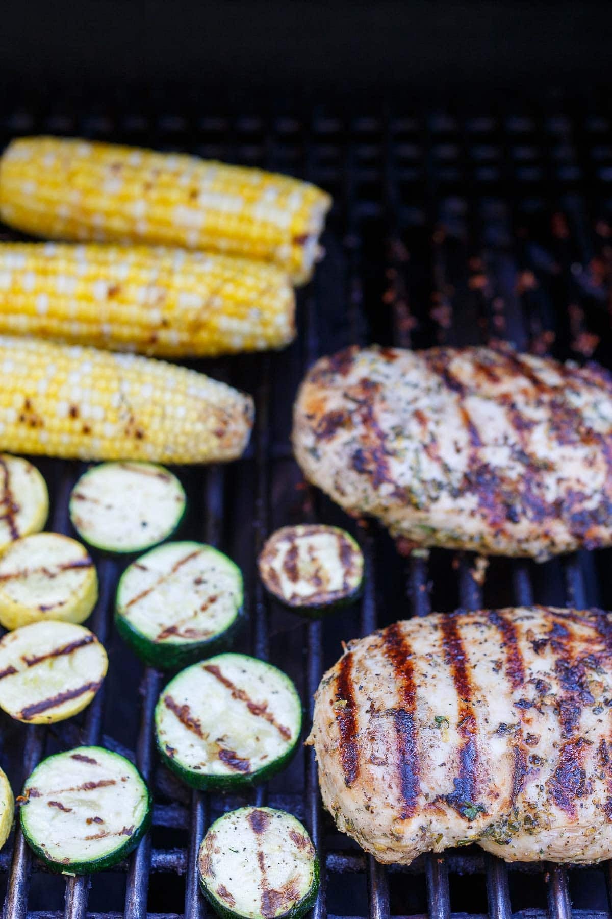 Mediterranean grilled chicken on the grill with grilled corn and sliced zucchini and yellow squash.