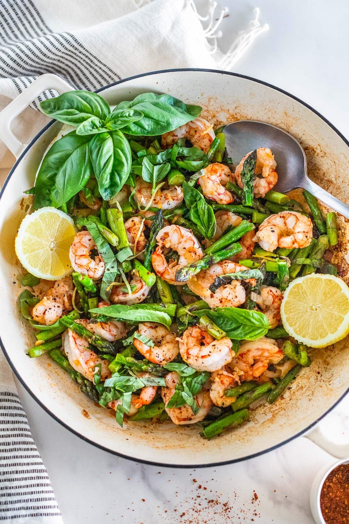 Skillet with one pan shrimp and asparagus with basil, lemon halves, and a spoon.