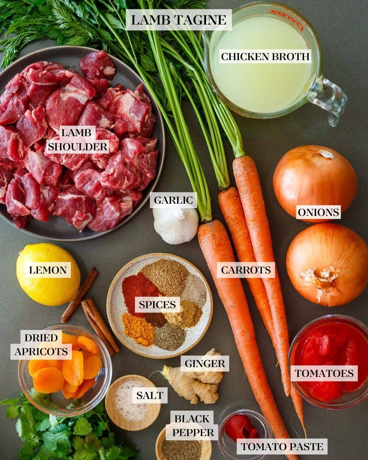 lamb tagine ingredients with plate of lamb shoulder, measuring cup of broth, two onions, three carrots, garlic bulb, small bowls of crushed tomatoes and tomato paste, plate of spices, fresh ginger, lemon, small bowl of dried apricots, two small bowls of salt and pepper, and fresh cilantro.