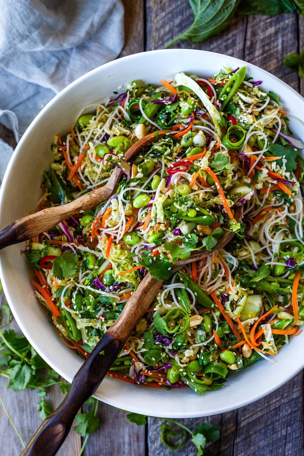 large bowl of Asian noodle salad with wood utensils, with shredded, colorful veggies and herbs, and sesame seeds and peanuts.