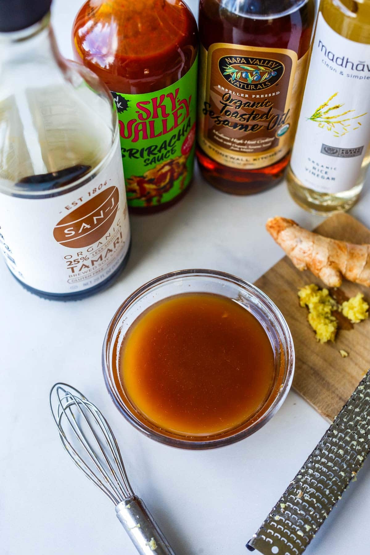 small bowl of Asian dressing next to a whisk, grated ginger, tamari, sriracha, toasted sesame oil, and rice vinegar.