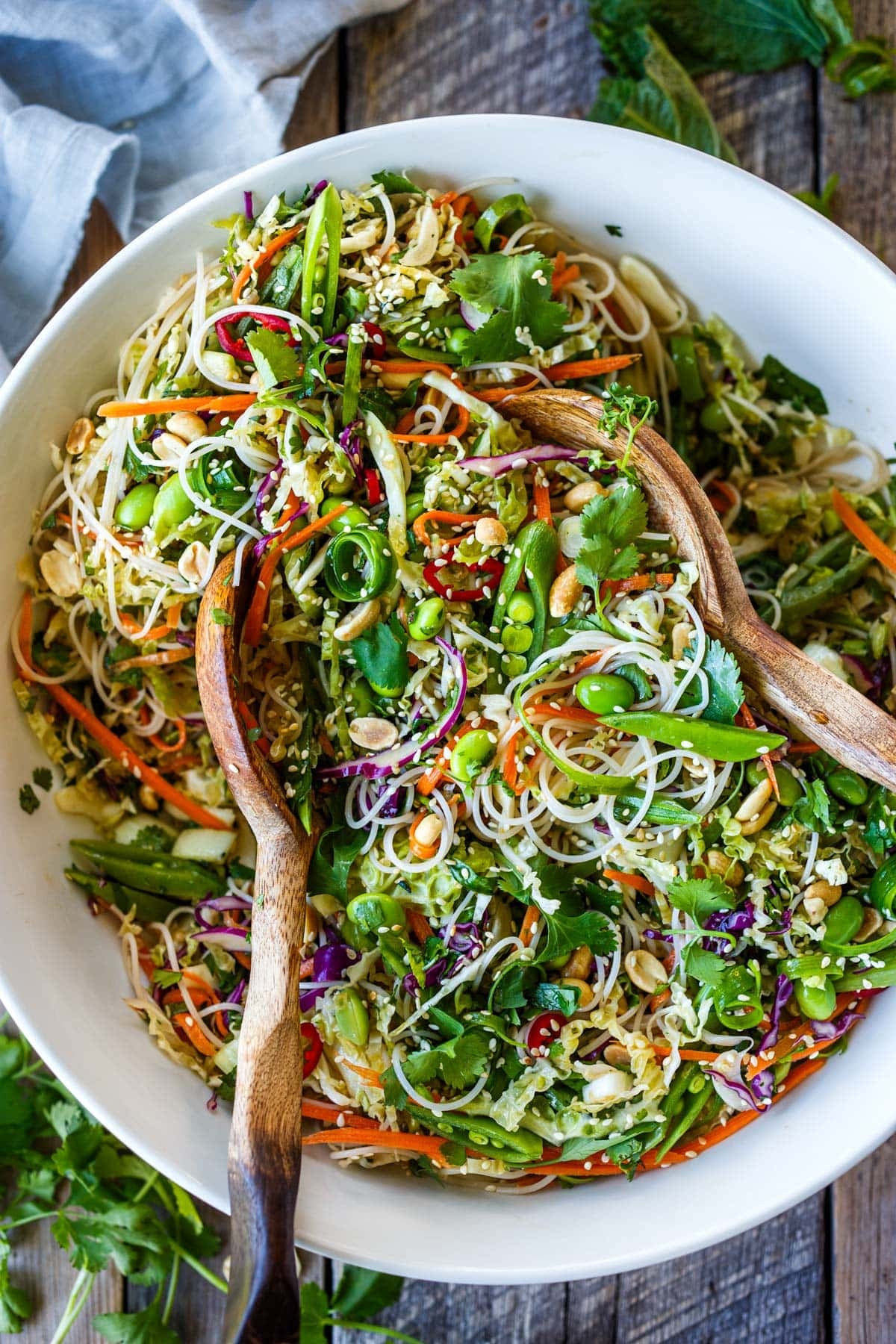 Asian noodle salad with edamame, snap peas, shredded carrots and cabbage, sesame seeds, and peanuts, in a bowl with wood utensils.