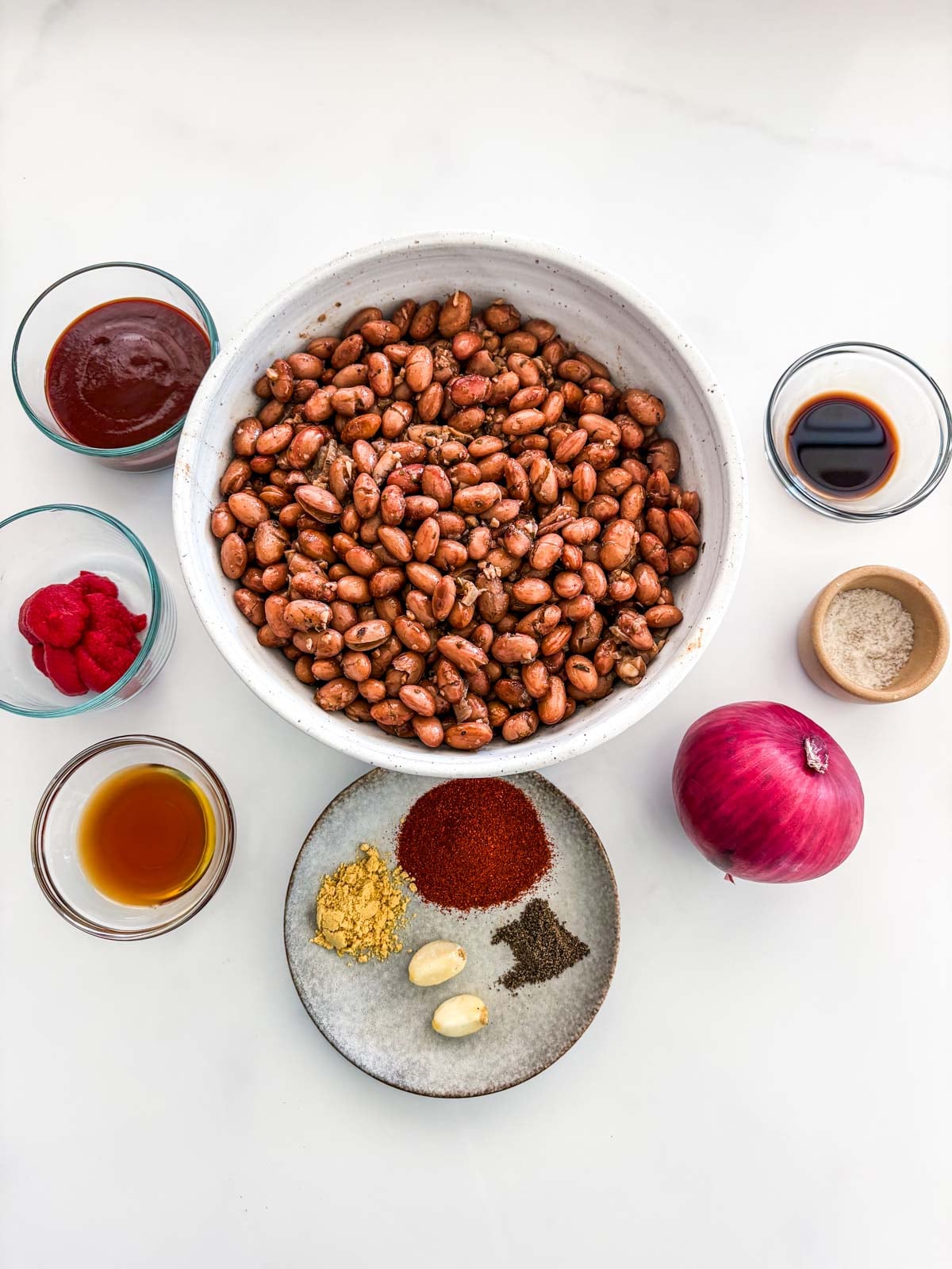 ingredients for baked beans - bbq sauce, tomato paste, maple syrup, spices and garlic cloves, red onion, salt and pepper, and worcestershire sauce surrounding a bowl of beans.