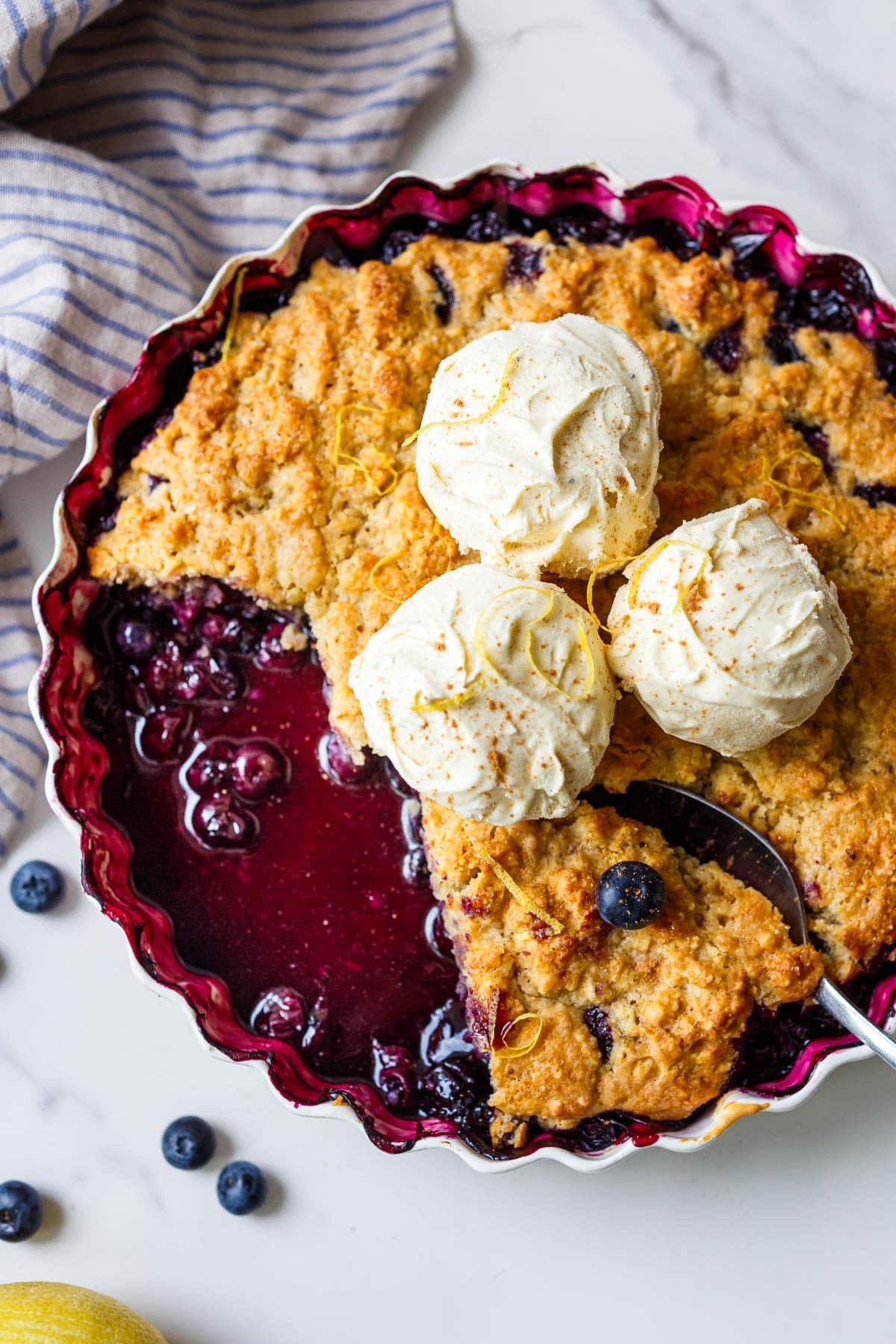 blueberry cobbler in a baking dish, topped with three scoops of ice cream and lemon zest, with a spoon removing a slice.