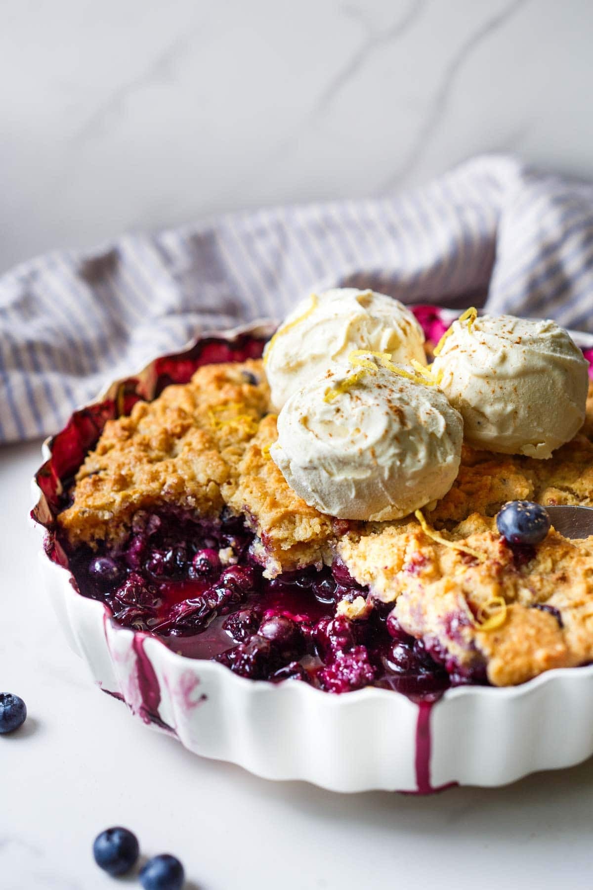 blueberry cobbler with pieces removed to show the juicy interior, topped with scoops of ice cream and lemon zest.