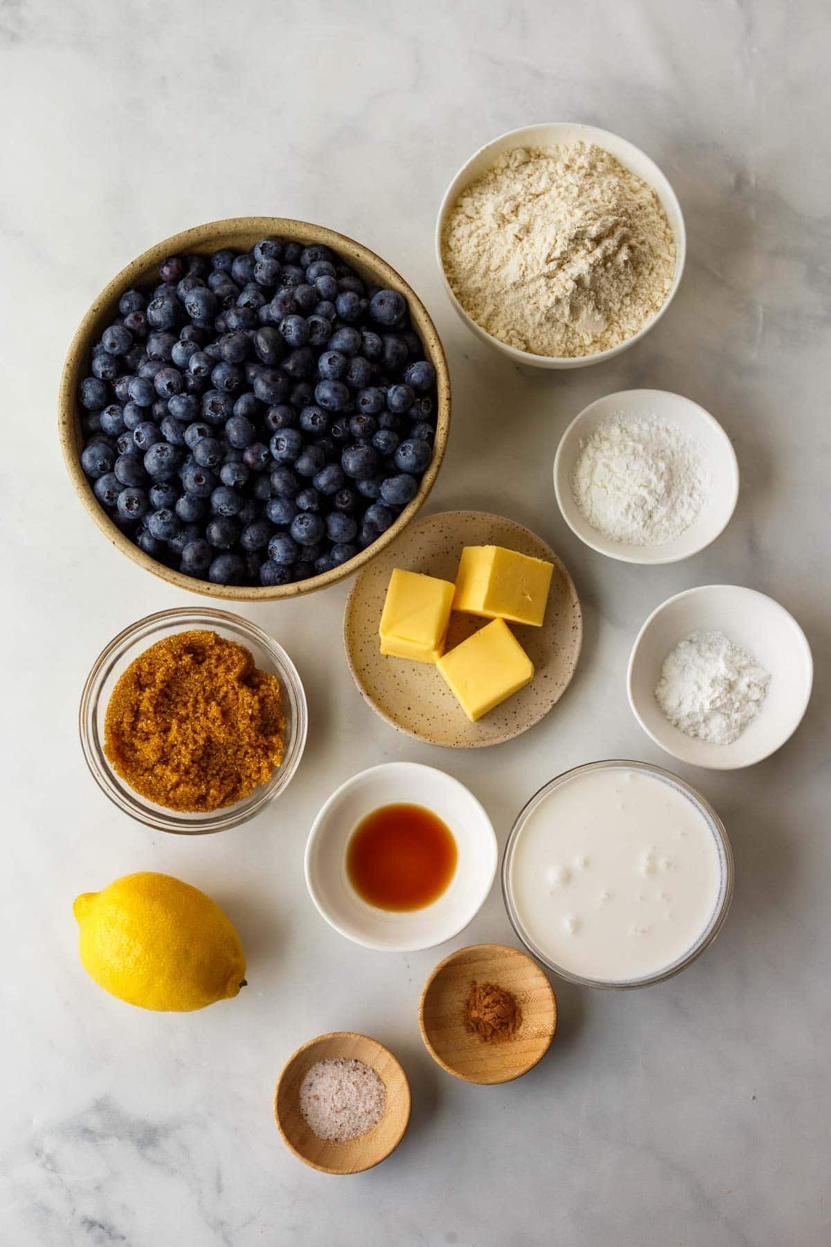 ingredients for blueberry cobbler - flour, cornstarch, baking soda, blueberries, unsalted butter, brown sugar, vanilla, milk, lemon, cinnamon, salt, arranged neatly in bowls.