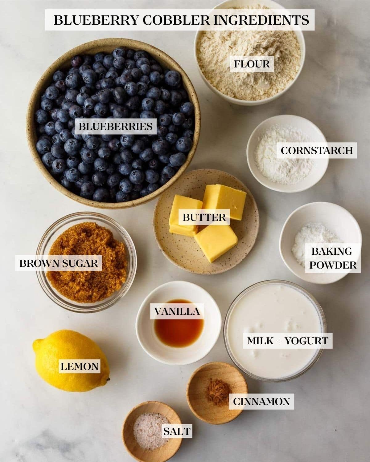 ingredients for blueberry cobbler - flour, cornstarch, baking soda, blueberries, unsalted butter, brown sugar, vanilla, milk, lemon, cinnamon, salt, arranged neatly in bowls. 