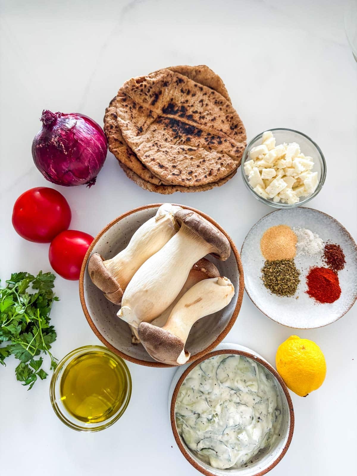 ingredients for Greek souvlaki pita - pita rounds, bowl of feta, plate of spices, red onion, tomatoes, bowl of king oyster mushrooms, fresh parsley, lemon, bowl of olive oil, and bowl of tzatziki.