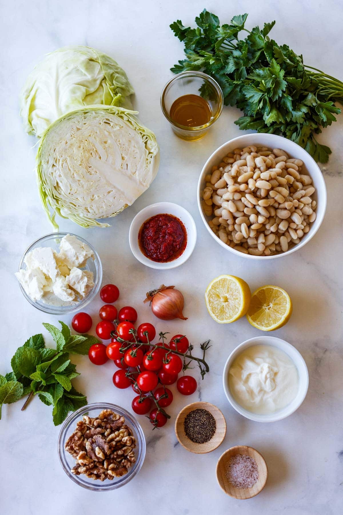 ingredients for grilled cabbage - parsley, olive oil, cabbage, white beans, harissa, feta, shallot, lemon, cherry tomatoes, Greek yogurt, mint, walnuts, pepper, and salt.