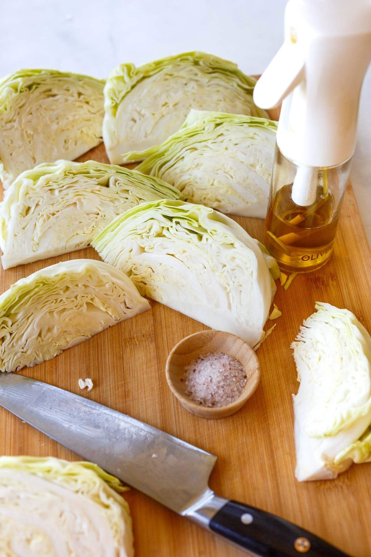 cabbage wedges on wood cutting board with pinch bowl of salt, sharp knife, and olive oil spray bottle.