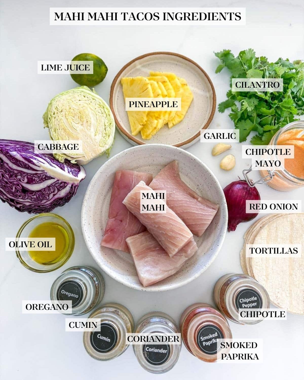 ingredients neatly arranged to make mahi mahi tacos - lime, cabbage, pineapple, cilantro, garlic cloves, chipotle mayo, red onion, tortillas, mahi mahi, oil, and spice jars.