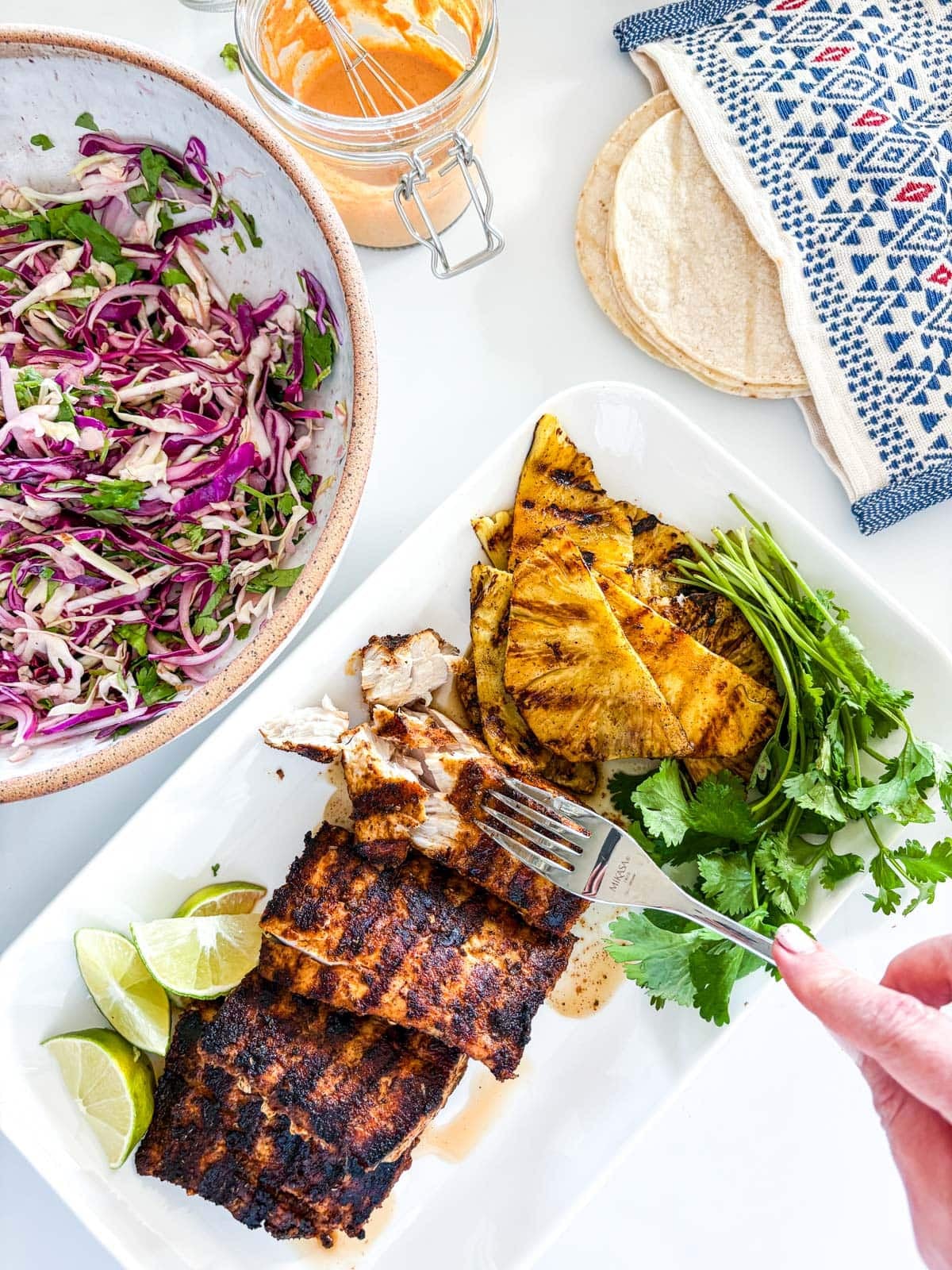 assembling mahi mahi tacos with jar of chipotle mayo, tortillas, cabbage slaw, cilantro, grilled pineapple, grilled mahi mahi, and lime wedges.