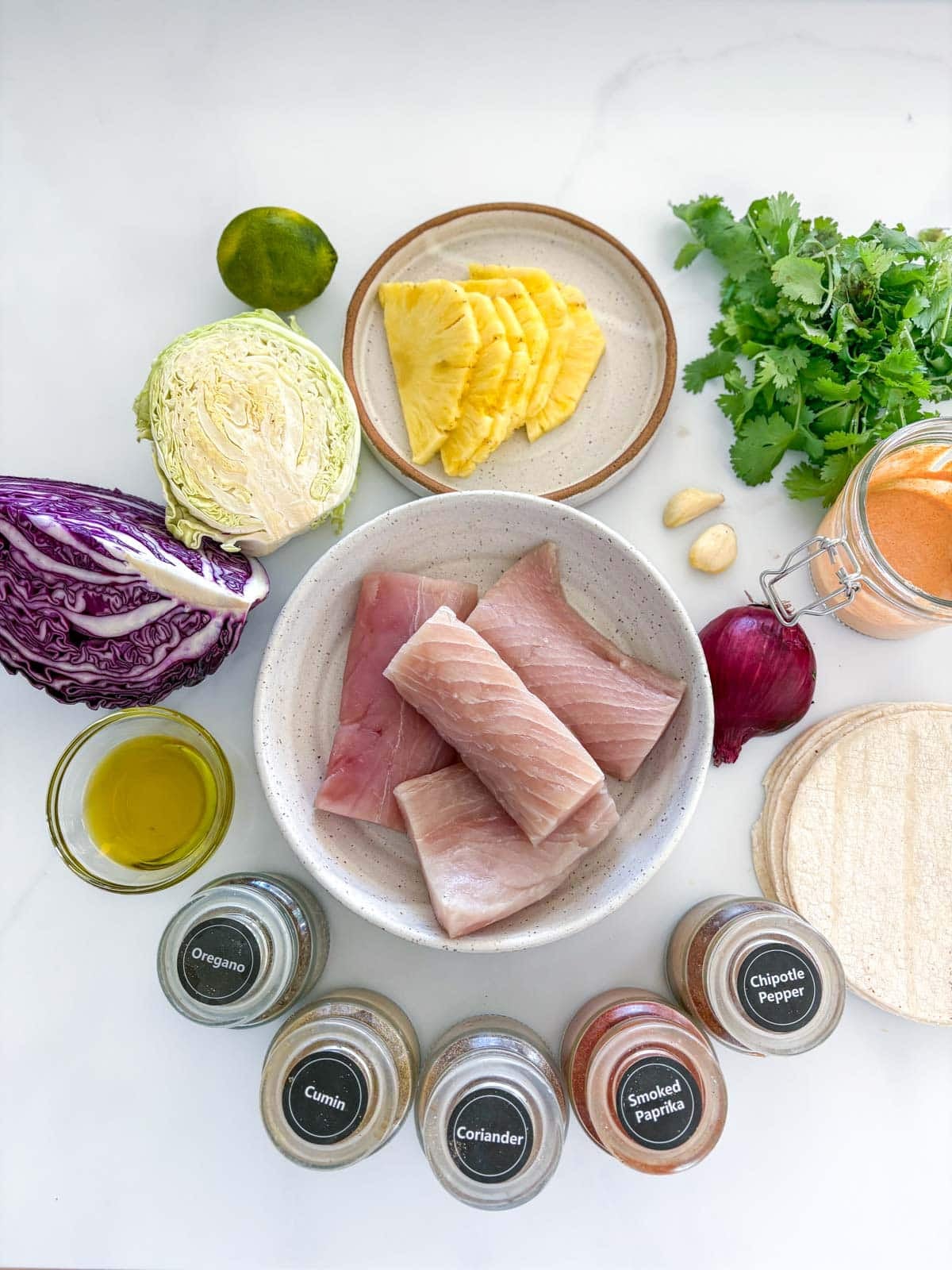 ingredients neatly arranged to make mahi mahi tacos - lime, cabbage, pineapple, cilantro, garlic cloves, chipotle mayo, red onion, tortillas, mahi mahi, oil, and spice jars.