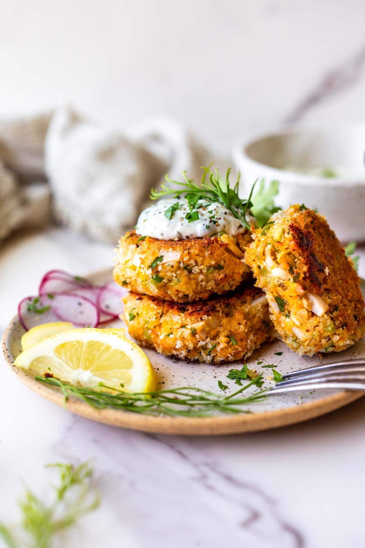 plate with three vegetarian crab cakes topped with lemon dill sauce and fresh dill, next to lemon wedges, more fresh dill, and sliced radish.