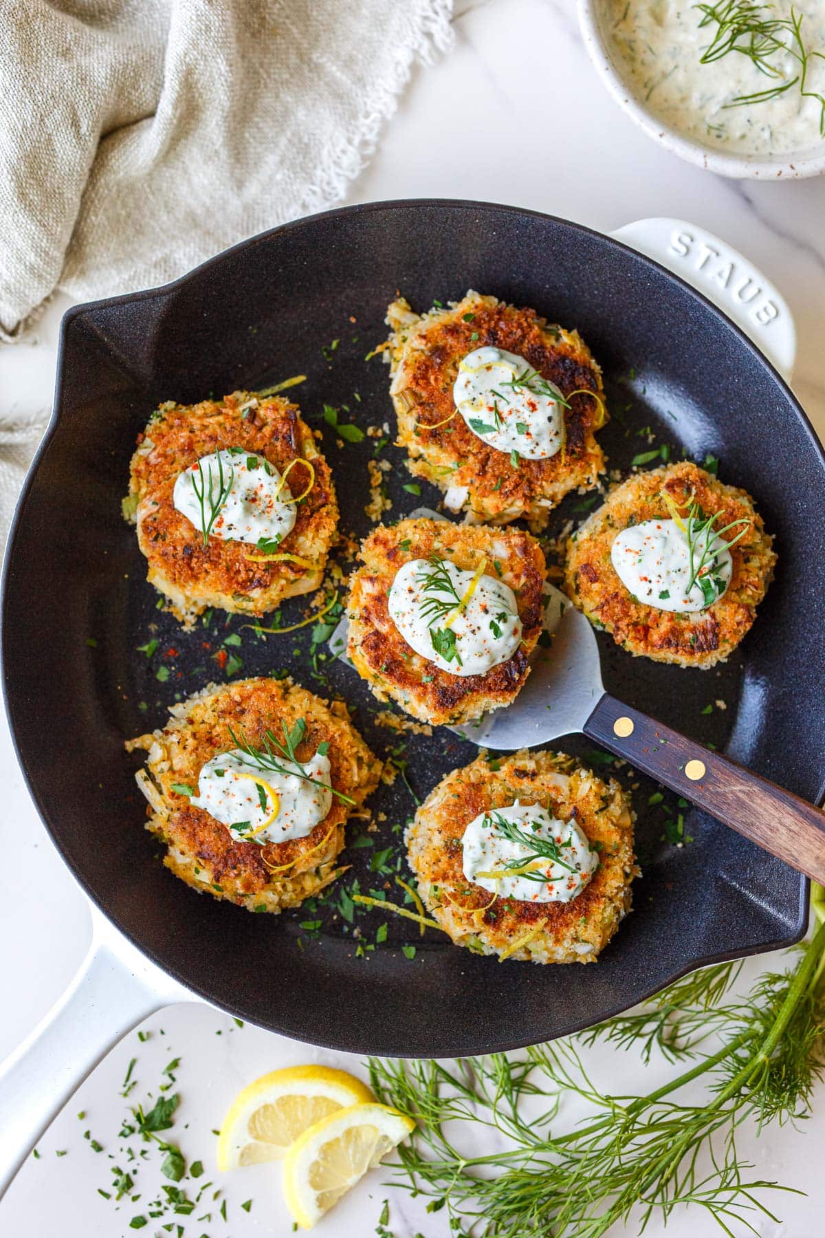 fried vegetarian crab cakes in a pan with dollops of lemon dill sauce, herbs, and lemon zest.