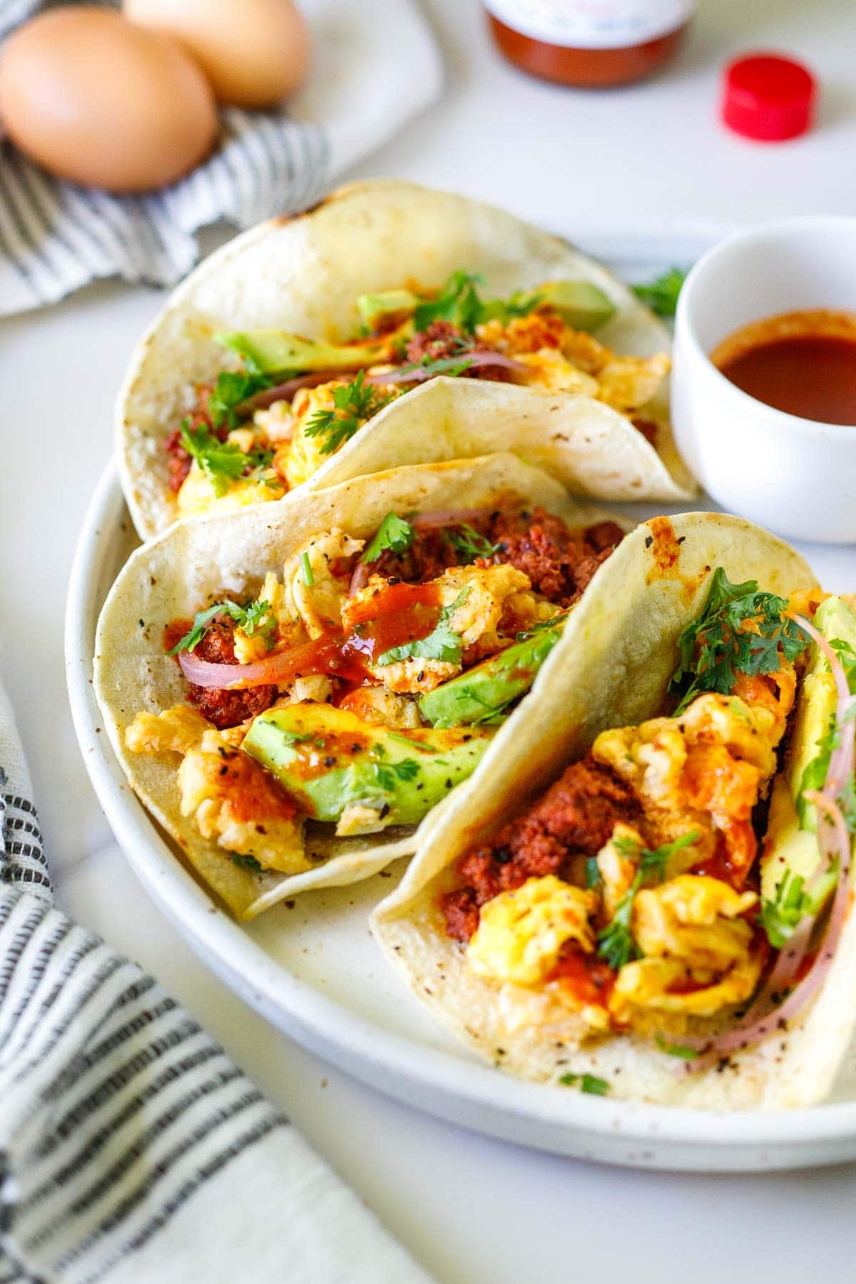 three breakfast tacos with eggs, chorizo, avocado, pickled onion, cilantro, and hot sauce on a plate with a small bowl of hot sauce.