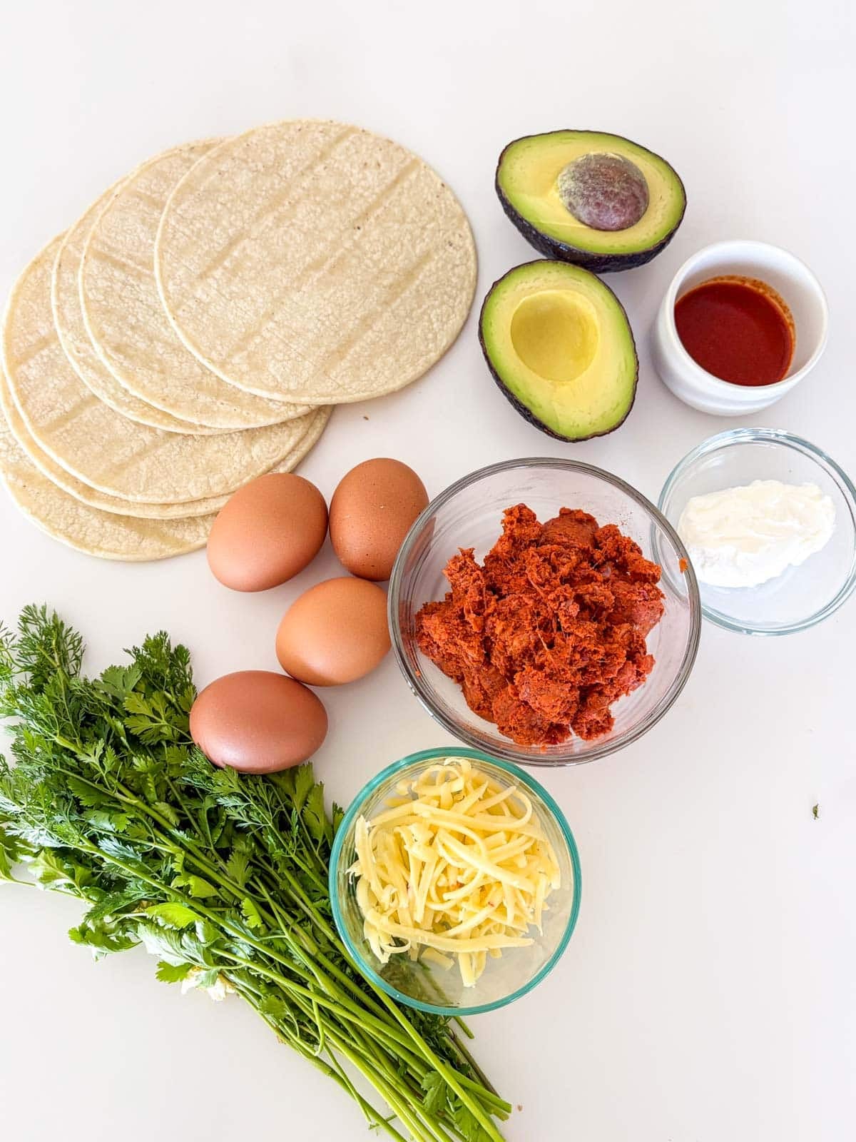 ingredients for breakfast tacos laid out - tortillas, avocado, hot sauce in a bowl, sour cream in a bowl, chorizo in a bowl, eggs, grated cheese in a bowl, and a bunch of fresh herbs.