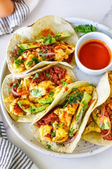 Breakfast tacos with chorizo and avocado, on a white dish wtih hot sauce.