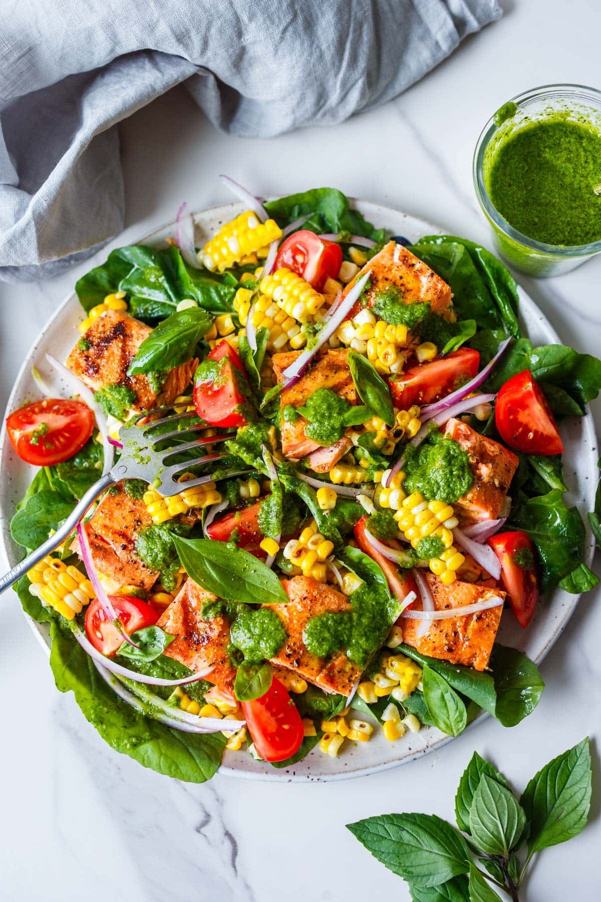 grilled salmon salad on a plate with tomato, red onion, and grilled corn, drizzled with basil vinaigrette and garnished with fresh basil.