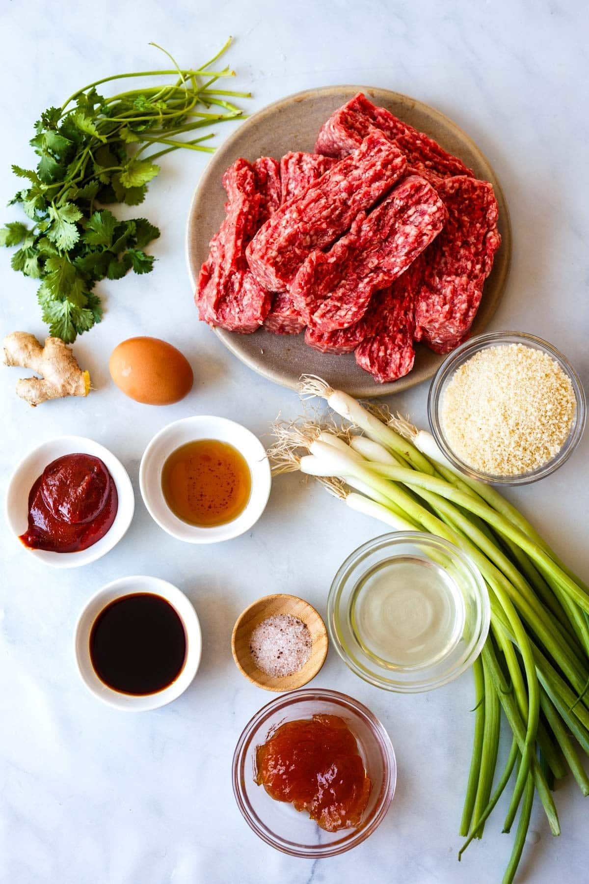 ingredients for Korean meatballs - plate of ground beef, fresh herbs, fresh ginger, egg, bowl of breadcrumbs, more small bowls of sauces, pinch bowl of salt, and bunch of green onions.