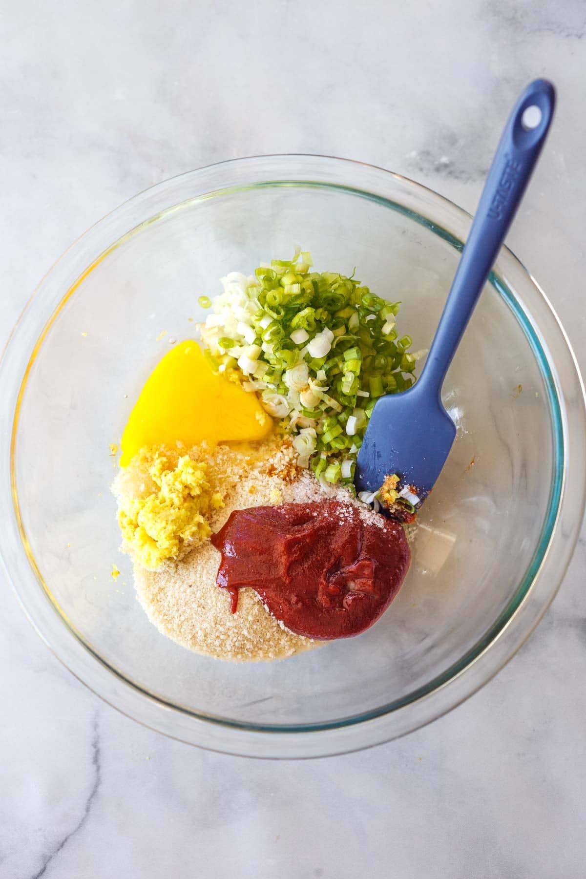 glass mixing bowl with the ingredients to make Korean meatballs - scallions, egg, ginger, breadcrumbs, and gochujang, with silicone spatula.
