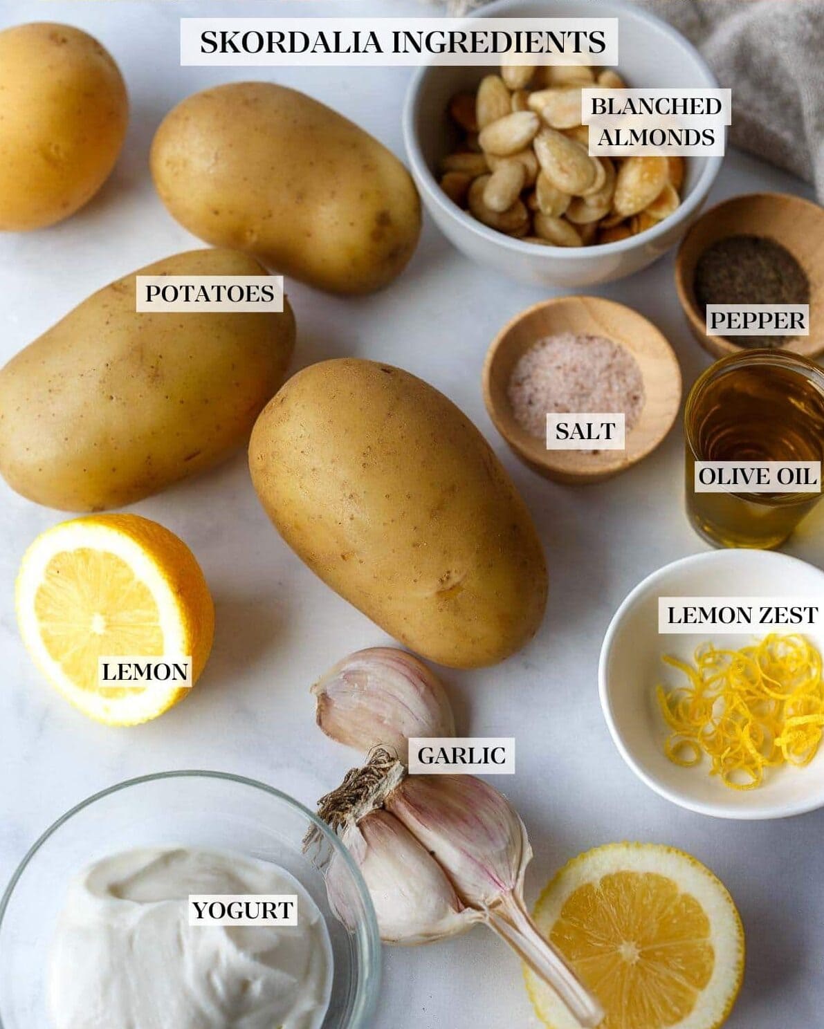skordalia ingredients - yellow potatoes, bowl of blanched almonds, salt and pepper, olive oil, bowl of lemon zest, garlic cloves, lemon halves, and bowl of yogurt.
