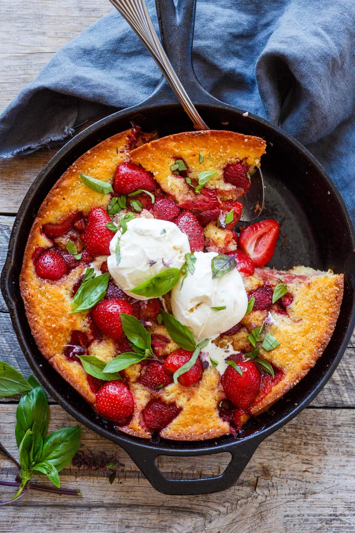 strawberry cobbler in a cast iron skillet with a piece removed, topped with two scoops of vanilla ice cream and fresh basil.