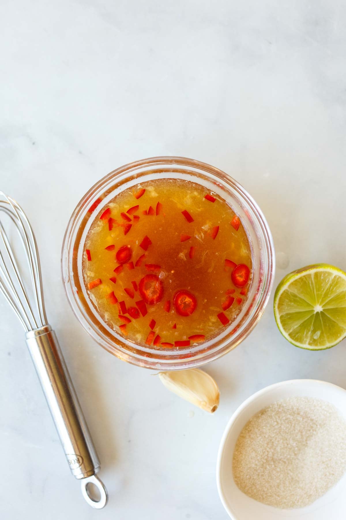 nuoc cham in a glass bowl next to lime, garlic clove, bowl of sugar, and a whisk.