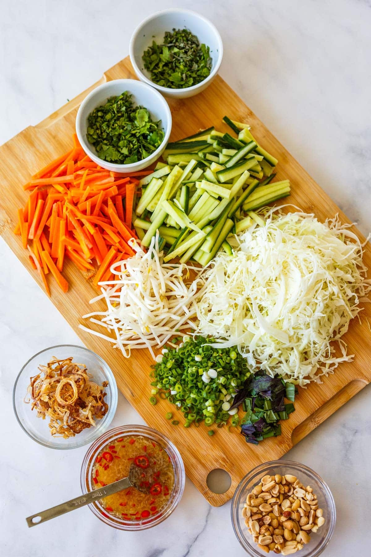 ingredients for Vietnamese vermicelli salad prepped and arranged on wood board - herbs, carrots, cucumbers, bean sprouts, shredded cabbage, scallions, crispy shallots, nuoc cham, and peanuts.