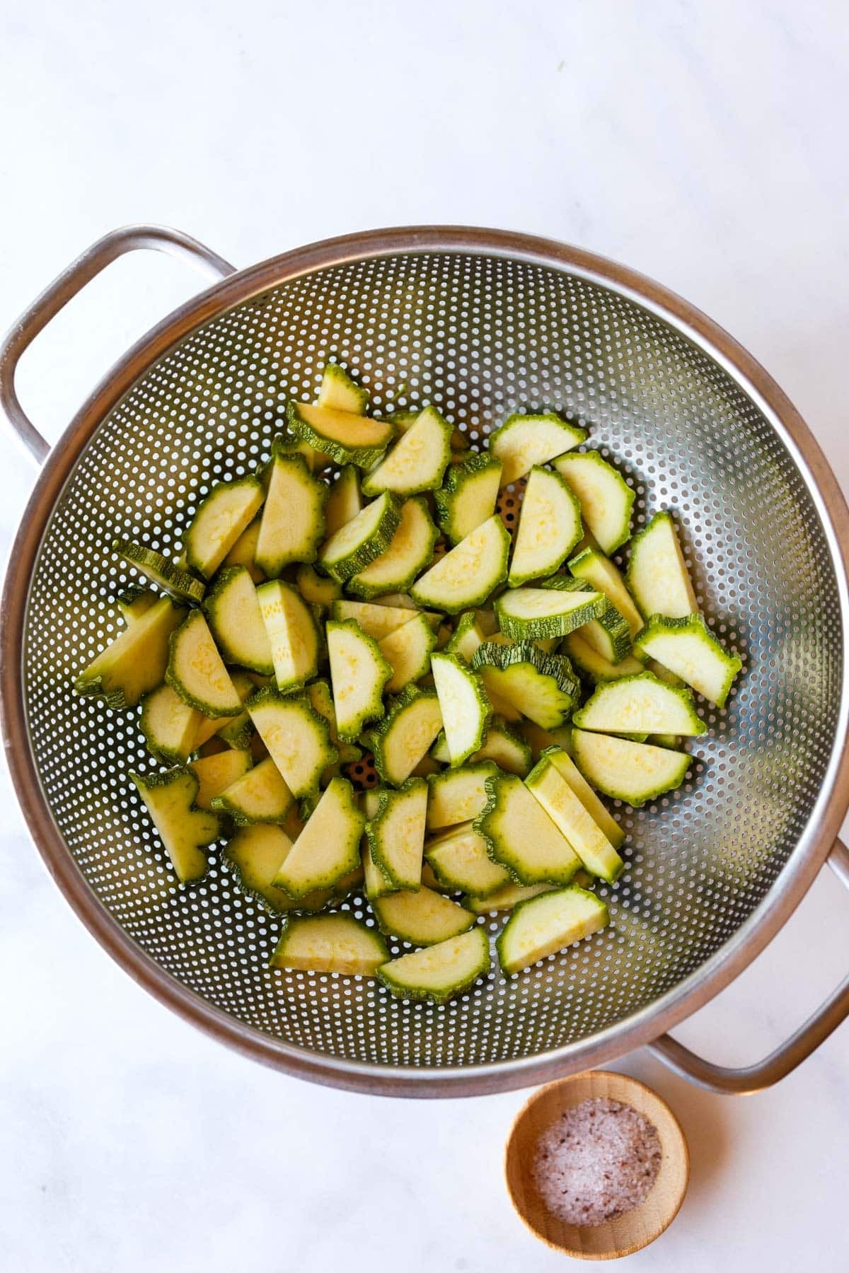 half moon zucchini slices in a colander with pinch bowl of salt.