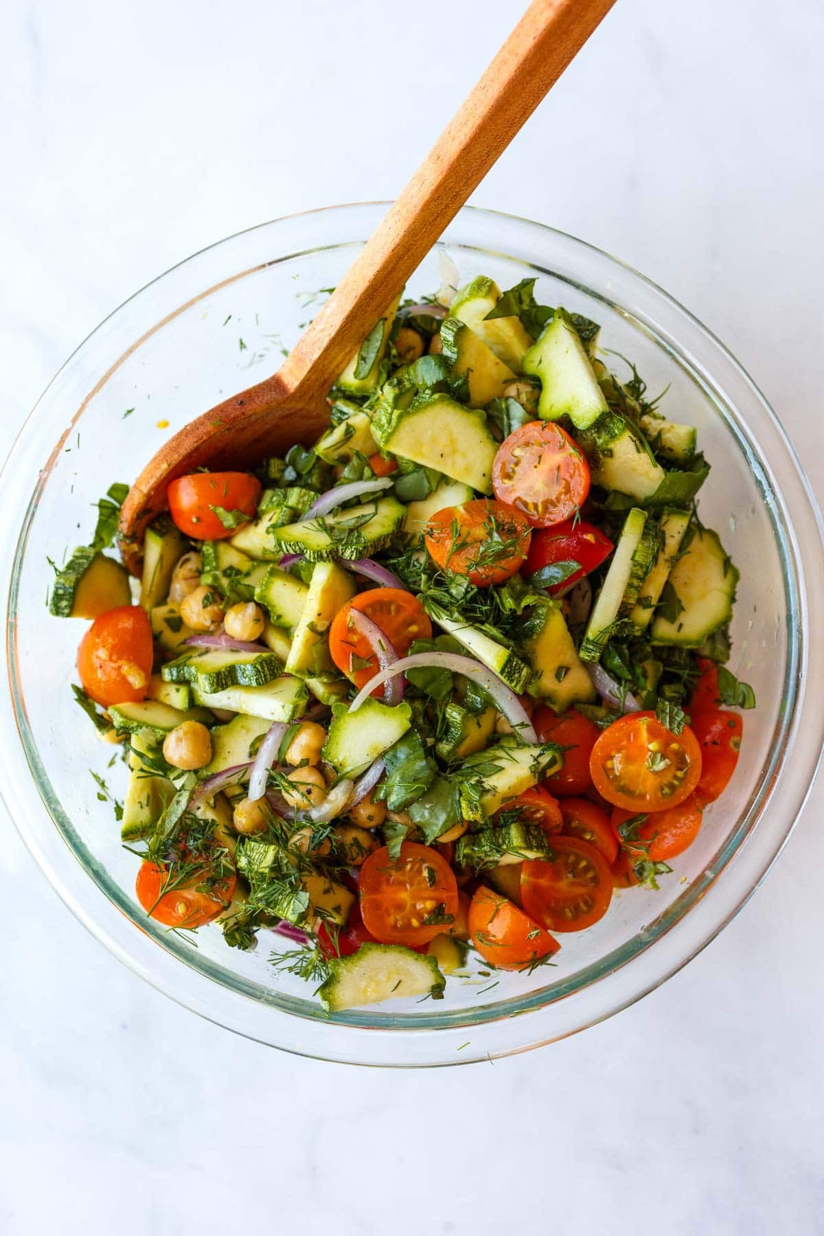 cherry tomatoes, sliced zucchini, thinly sliced red onion, chickpeas, and chopped fresh herbs in a glass mixing bowl to make zucchini salad with a wood spoon.