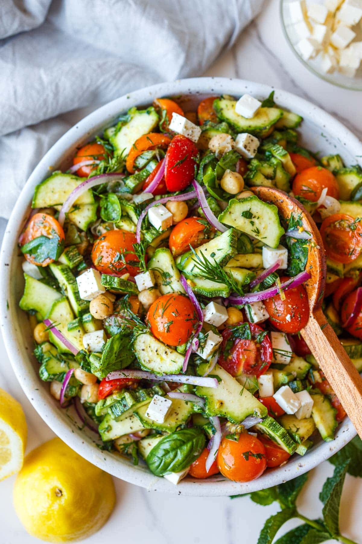 zucchini salad with herbs, red onion, cherry tomatoes, chickpeas, and fresh herbs in a salad bowl with wood serving utensil.