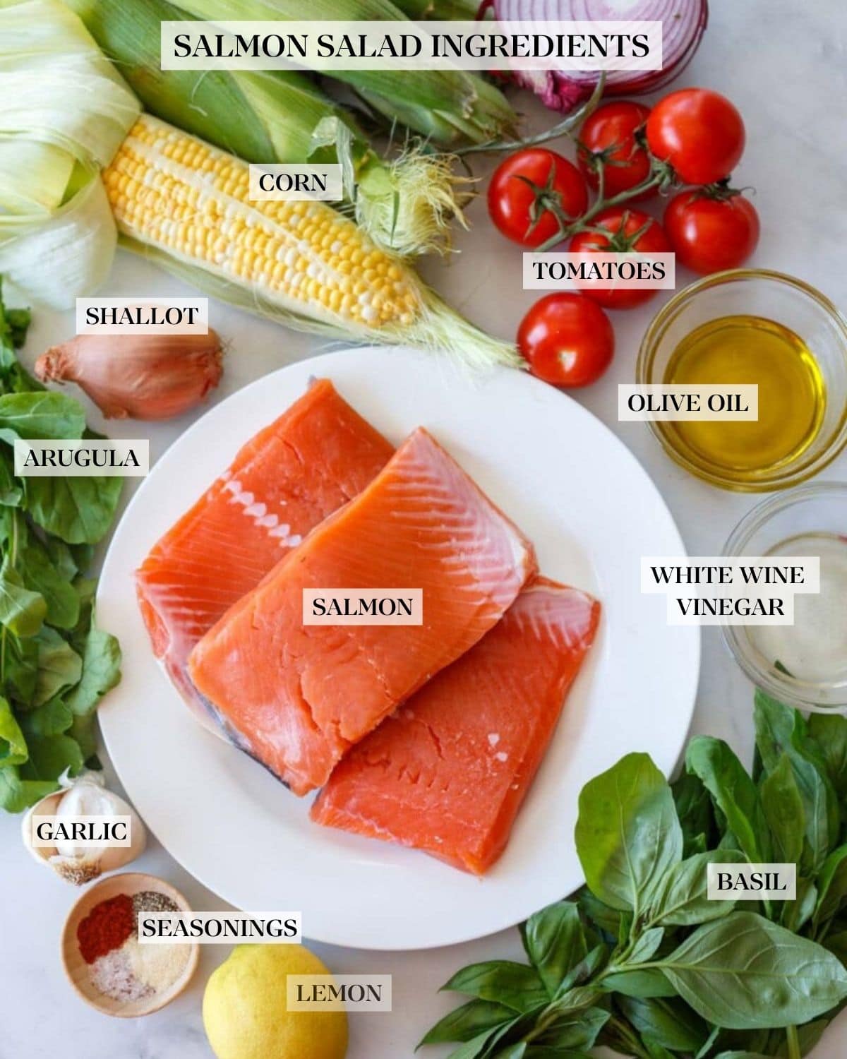 salmon salad ingredients laid out - ears of corn, red onion, vine tomatoes, bowl of olive oil, shallot, basil bunch, plate of salmon filets, vinegar in a bowl, garlic bulb, pinch bowl of dry rub spices, and lemon.