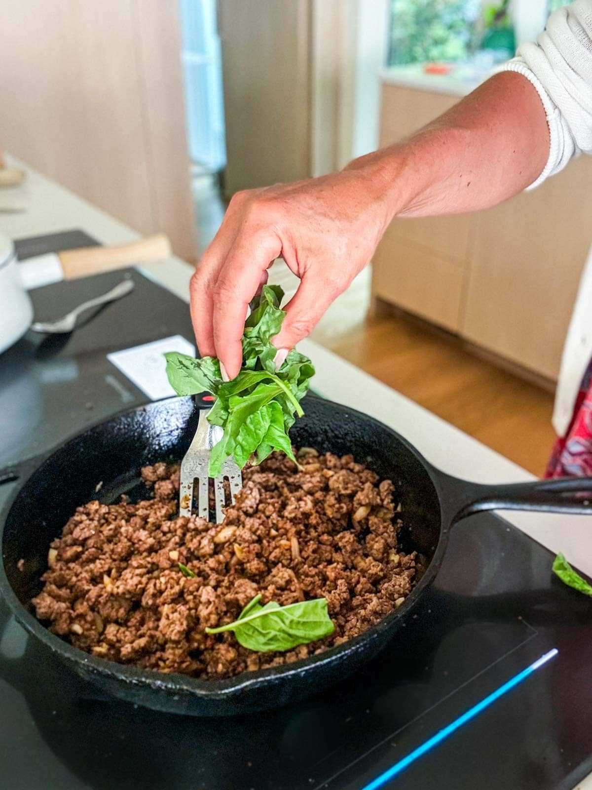 hand adding a handful of fresh basil to the cast iron skillet of ground beef to make basil beef. 