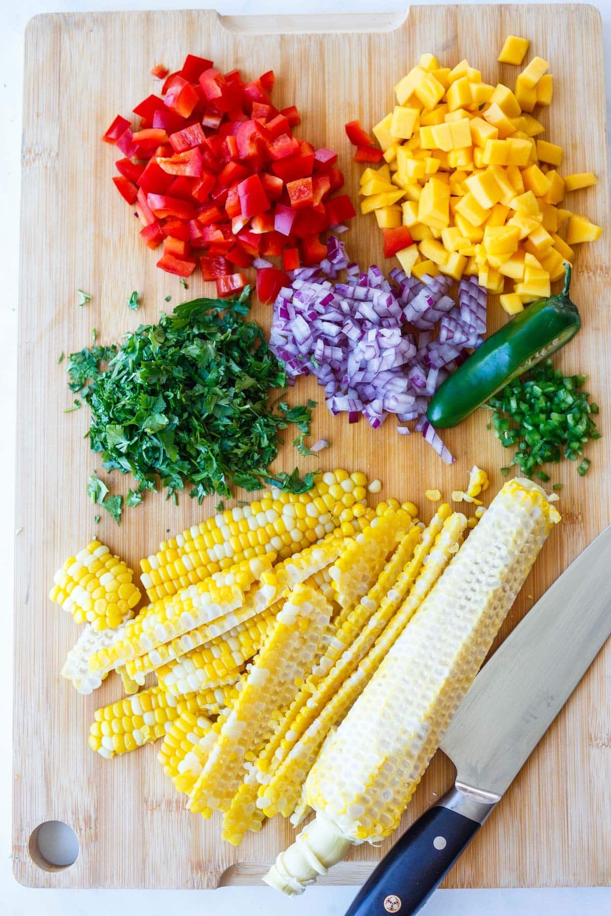 chopped veggies for black bean salad on a wood board with a sharp knife - red bell pepper, mango, red onion, cilantro, jalapeño, and corn.