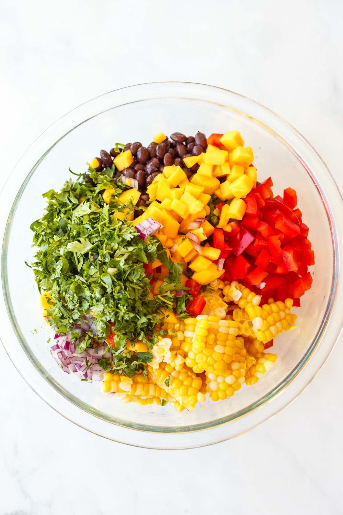 glass bowl with each component added to make black bean salad - black beans, mango, red bell pepper, corn, red onion, and fresh cilantro.