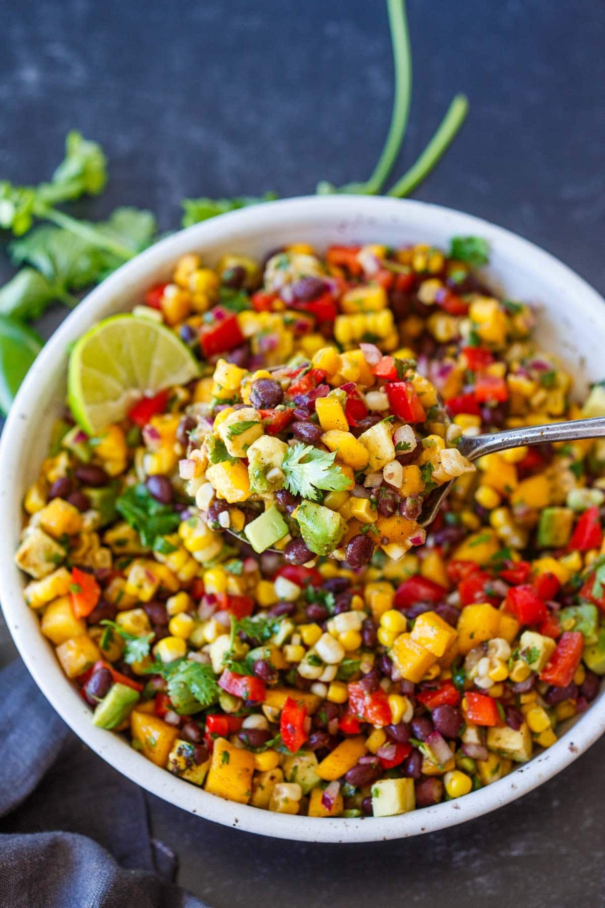 black bean salad with mango, corn, avocado, and red bell pepper in a bowl with a lime wedge, fresh cilantro, and a spoon lifting a spoonful up.
