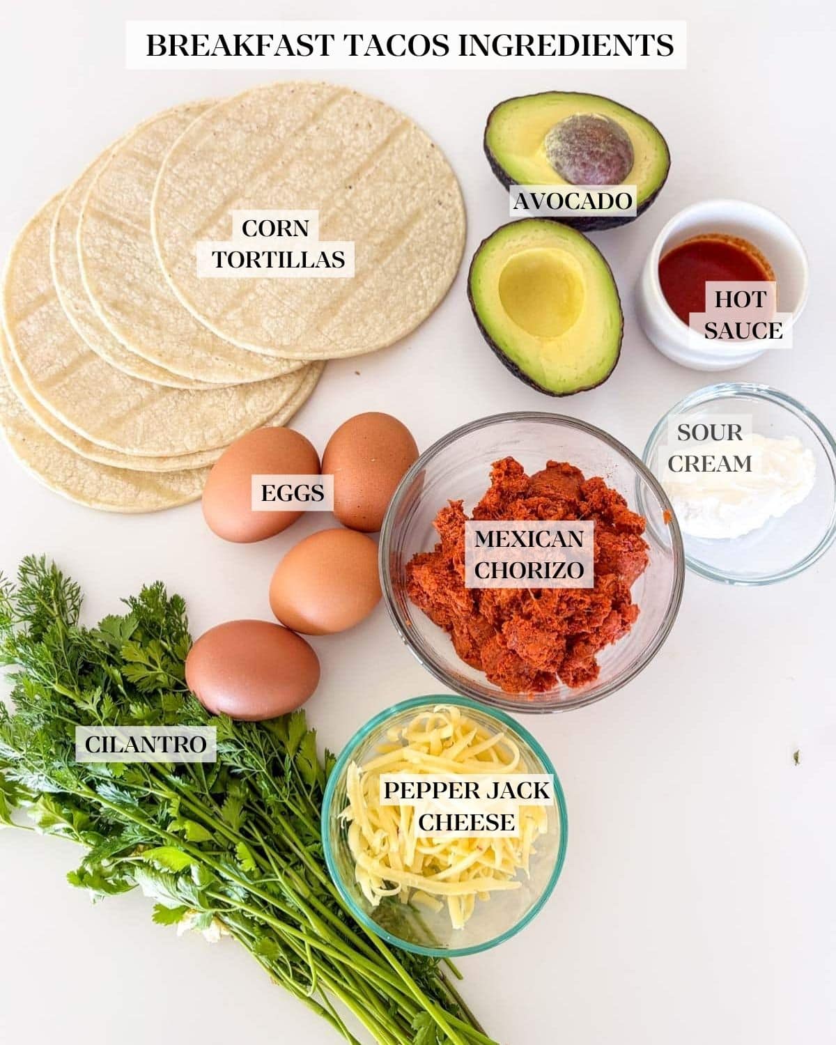ingredients for breakfast tacos laid out - tortillas, avocado, hot sauce in a bowl, sour cream in a bowl, chorizo in a bowl, eggs, grated cheese in a bowl, and a bunch of fresh herbs.