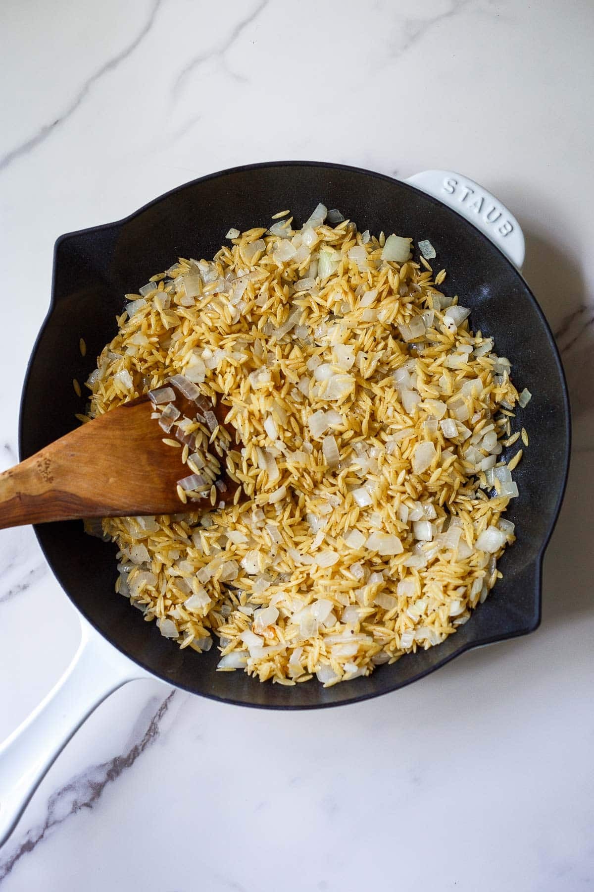 Orzo and onions in a skillet with a wood spatula.