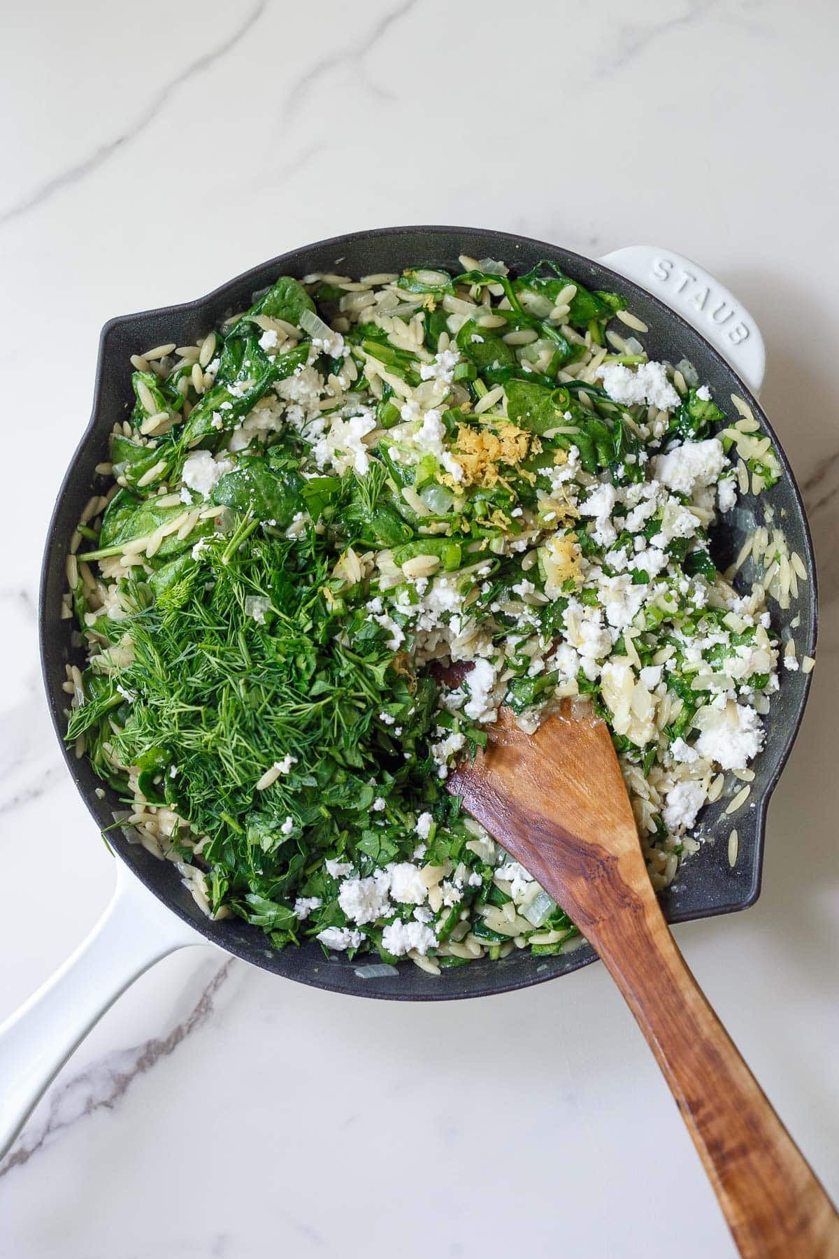Finishing the dish- skillet with orzo, fresh herbs, lemon zest, and feta with a wood spatula, seasoning the greek orzo.