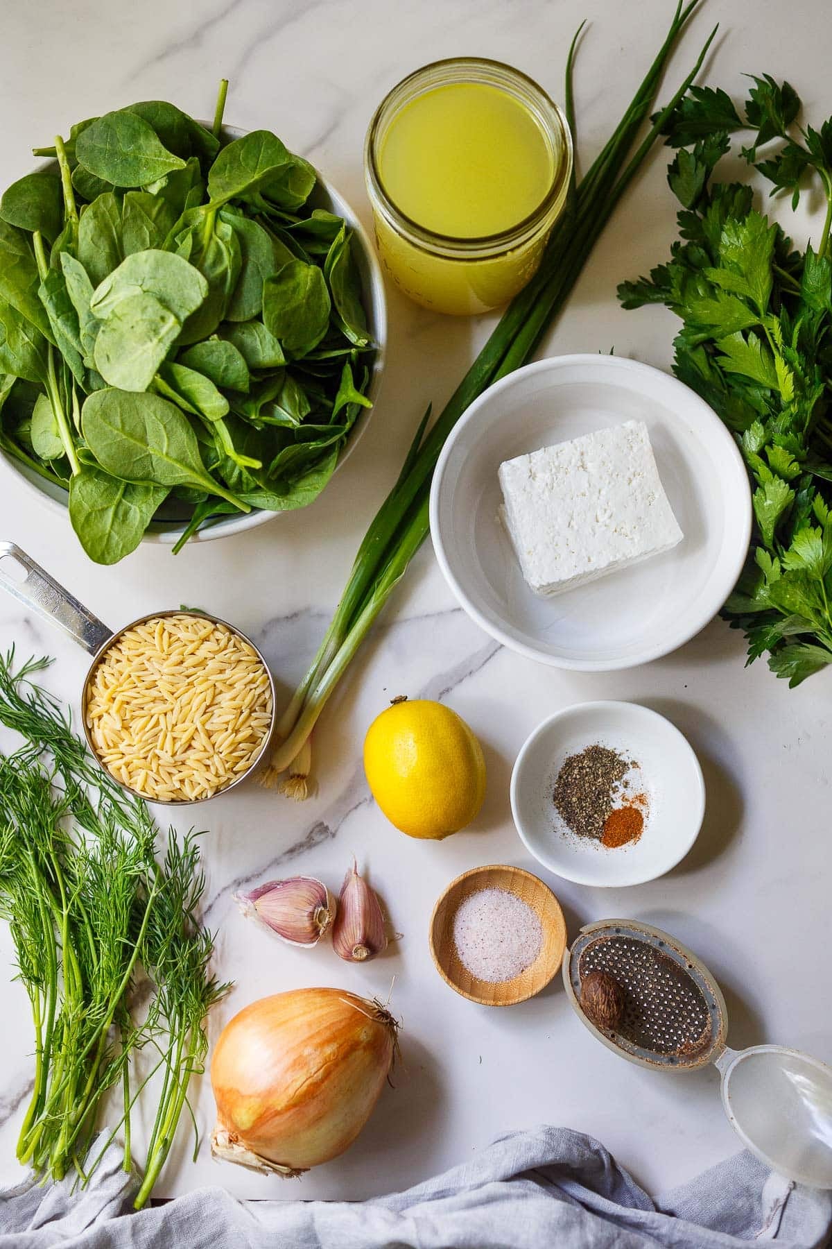 ingredients neatly arranged for one-pan Greek Orzo - broth, bowl of baby spinach, parsley bunch, block of feta, green onions, scoop of orzo, lemon, garlic cloves, pinch bowls with pepper, cayenne, and salt, fresh nutmeg, onion, and dill bunch.