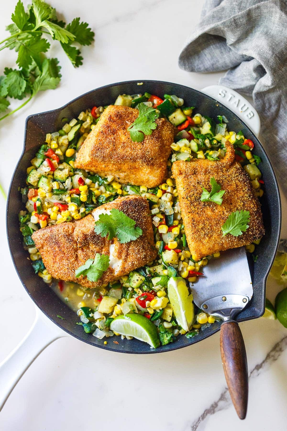 cornmeal-crusted halibut in a skillet on top of summer succotash, garnished with fresh cilantro and lime wedges.
