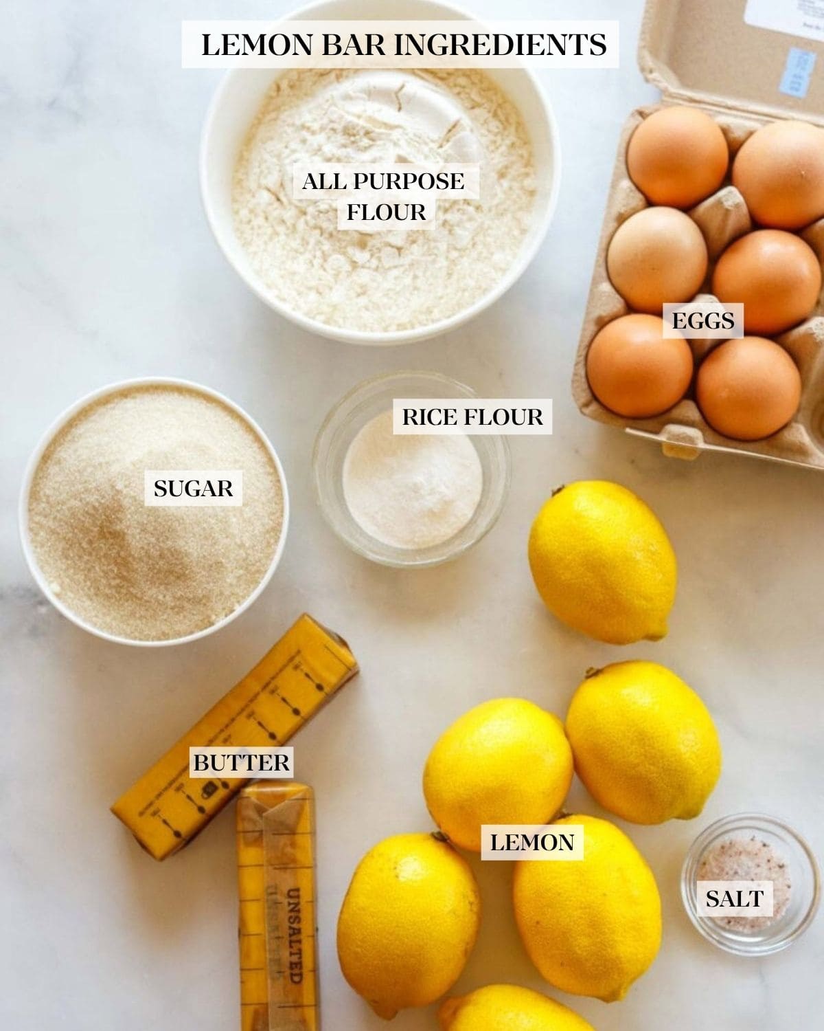 bowl of flour, carton of eggs, sugar, rice flour, lemons, butter sticks, and pinch bowl with pink salt - ingredients to make lemon bars.