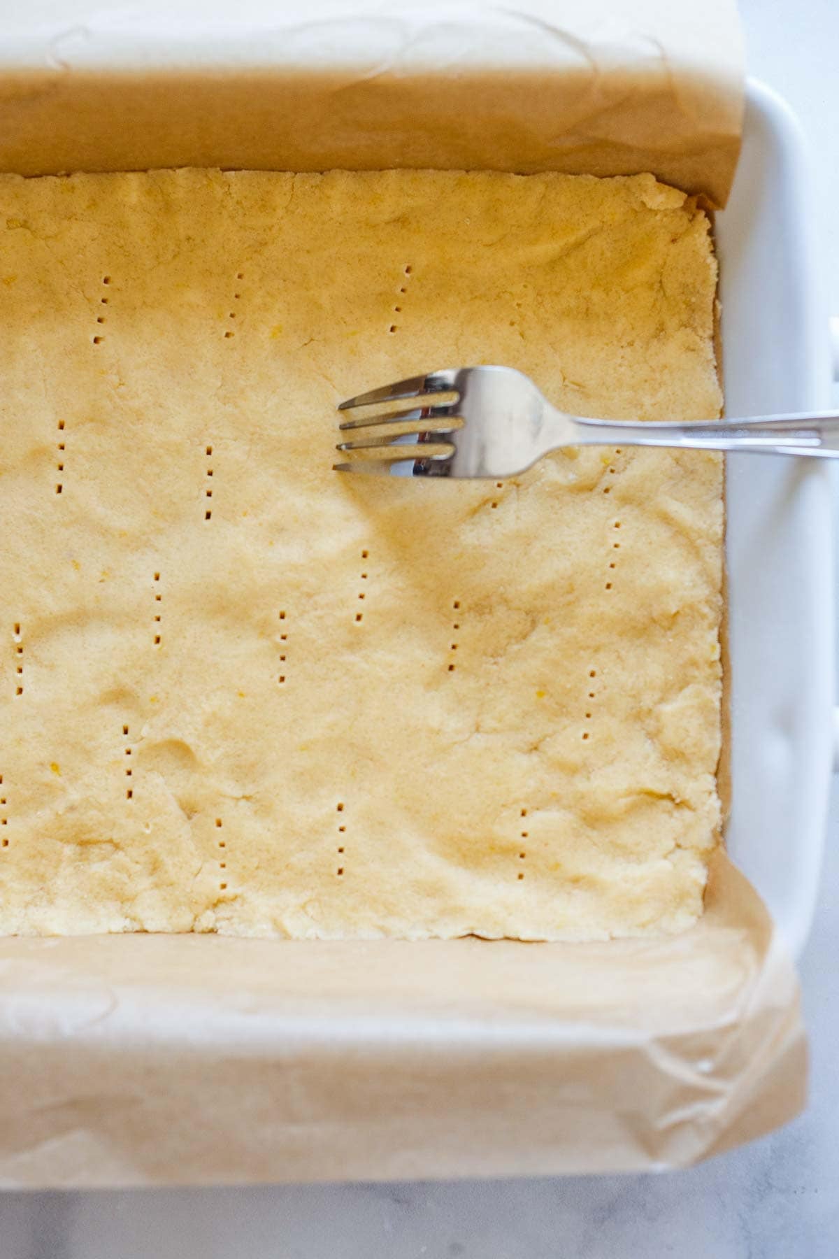 fork making pricks in shortbread crust on a parchment-lined baking tray.